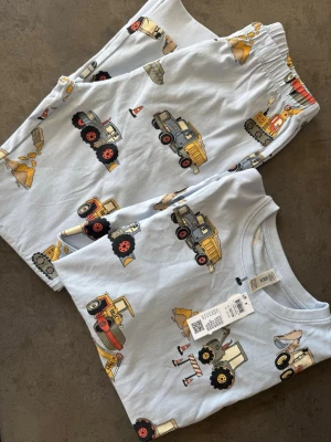 Ljusblå pyjamas med byggfordon - Säljer en ljusblå pyjamas med tryck av olika byggfordon som grävmaskiner, lastbilar och traktorer. Setet består av långärmad tröja och byxor med resår i midjan. Materialet är mjukt och skönt, perfekt för att chilla hemma eller sova i.