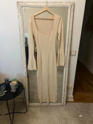 Beige ribbad långklänning Adoore - Trendig beige långklänning från Adoore med ribbad struktur och v-ringad hals. Klänningen har långa ärmar med lätt utsvängda ärmslut och en kroppsnära passform som framhäver siluetten. Perfekt för dig som gillar stilrena och moderna plagg.
