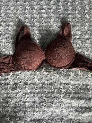 Brun spets-bh med vaddering - Snygg brun bh med vadderade kupor och spetsdetaljer framtill. Bh:n har justerbara axelband och hakknäppning bak. Materialet är en mix av spets och slät textil som ger en feminin look.