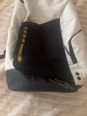 Svartvit ryggsäck från Nike - Snygg svartvit och guld ryggsäck från Nike med gulddetaljer och logga. Ryggsäcken har flera fack med dragkedja, justerbara axelband och sportig design. Perfekt för skola eller träning och rymmer allt du behöver. Materialet är slitstark syntet.
