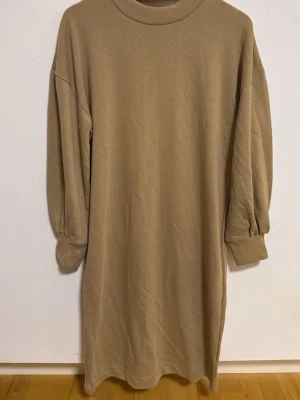 Beige långärmad klänning - En enkel och stilren beige klänning med lång ärm och ribbad hög krage. Klänningen har en loose passform och är gjord i mjukt bomullsmaterial, perfekt för en avslappnad look.