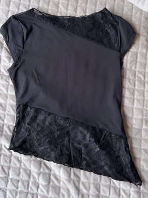 Svart topp med spetsdetaljer - Snygg svart topp med asymmetrisk design och spetsdetaljer både upptill och nedtill. Toppen har korta ärmar och en lätt loose passform. Perfekt för dig som gillar en stilren look med lite extra edge. Från SHEIN.