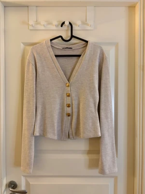 Beige kofta med guldfärgade knappar - Stilren beige kofta med v-ringning och fyra dekorativa guldfärgade knappar framtill. Modellen är något croppad med långa ärmar och mjukt, tunt stickat material. Köpt på Zara men aldrig använts