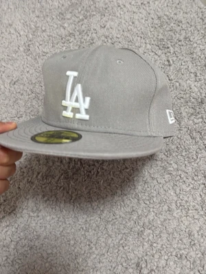Grå LA New Era 59FIFTY keps - Snygg grå keps från New Era 59FIFTY med LA-logga broderad i vitt framtill. Klassisk flat brim och strukturerad form. Märke broderat på sidan. Tillverkad i ett tåligt tygmaterial, perfekt för en streetwear-look.
