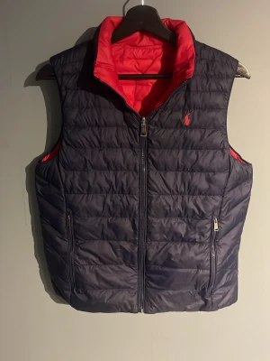 Vändbar Ralph Lauren dunväst i blå och rött - Snygg vändbar dunväst från kidsbrandstore med ena sidan blå och andra sidan röd. Västen har dragkedja framtill, två fickor med dragkedja och en liten logga på bröstet. Perfekt för dig som vill variera din stil och hålla värmen utan att tumma på looken. Passar även till vår och sommar.