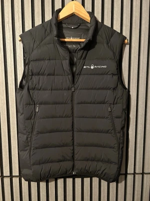 Svart dunväst från Sail Racing - Stilren svart dunväst från Sail Racing med diskret logga på bröstet och en på ryggen. Västen har två dragkedjeförsedda fickor och quiltad design som ger en sportig look. Perfekt för lager-på-lager och håller dig varm under kyliga dagar. Har dock något vitt märke under bröstet annars e de bra skick 