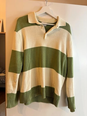Randig rugbytröja i grönt och beige - Snygg rugbyinspirerad tröja med breda gröna och beige ränder. Tröjan har krage, knappar vid halsen och ribbade muddar vid ärmslut och nederkant. Perfekt för en avslappnad och trendig look.
