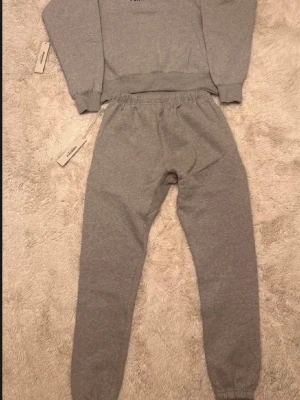 Grå Essentials tracksuit - Säljer ett grått loungewear set från Essentials med matchande hoodie och byxor. Hoodien har magficka och dragsko i huvan, byxorna har resår i midjan och muddar vid benslut. Båda plaggen har Essentials-logga tryckt framtill. Perfekt för chill dagar hemma.