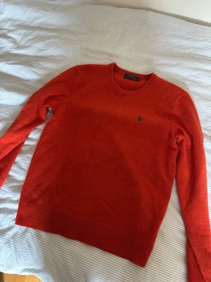 Röd stickad tröja från Polo Ralph Lauren - Snygg röd stickad tröja från Polo Ralph Lauren med rund halsringning och klassisk grön logga broderad på bröstet. Tröjan har ribbade muddar vid ärmslut och nederkant. Perfekt för dig som gillar stilrena och tidlösa plagg.