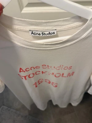Vit Acne Studios t-shirt - Vit t-shirt från Acne Studios med rött tryck på bröstet där det står 'Acne Studios STOCKHOLM 1996'. Klassisk rund hals och korta ärmar. Tillverkad i mjuk bomull för en skön känsla och avslappnad passform.