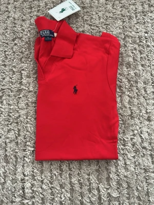 Röd pikétopp från Polo Ralph Lauren - Säljer en helt ny röd tröja/skjorta från Polo Ralph Lauren som är tillverkad av bomull med korta ärmar och krage. loggan är mörkblå och sitter på bröstet. tröjan är aldrig använd och taggen finns kvar.❤️