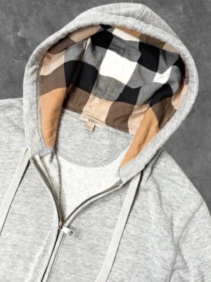 Burberry kofta - Säljer en grå zip hoodie från Burberry med klassiskt rutigt foder i huvan. Hoodien har dragsko i huvan, två fickor framtill och ribbade muddar vid ärmar och nederkant. Perfekt för en avslappnad och stilren look.