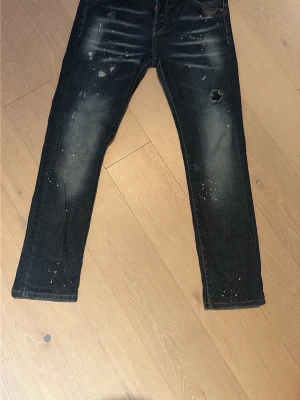 Mörkblå jeans från Dsquared2 - Säljer ett par mörkblå jeans från Dsquared2 med slitningar, färgstänk och distressed detaljer. Jeansen har raka ben och klassisk femficksmodell. Snygga kontrastsömmar och cool streetkänsla. Perfekta för dig som gillar en trendig och avslappnad stil.