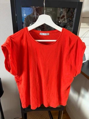Röd ribbad topp från Zara - Säljer en röd ribbad topp från Zara med rund halsringning och korta ärmar. Toppen har en loose passform och är tillverkad i mjukt material som känns skönt mot huden. Perfekt att styla med jeans eller kjol för en enkel look.