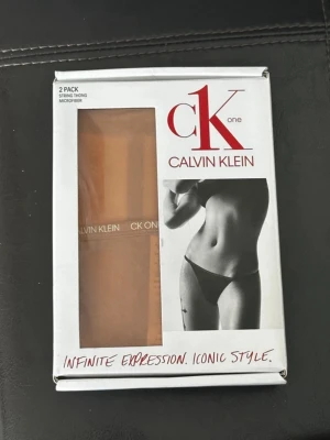Calvin Klein stringtrosor nude M. Oöppnad förpackning - 2-pack stringtrosor från Calvin Klein i nude färg, tillverkade i mjuk och stretchig mikrofiber. Klassisk CK One-design med logotyp i midjan. Perfekta för en diskret look under kläderna. Oöppnad förpackning!! Bild 2&3 är lånade.