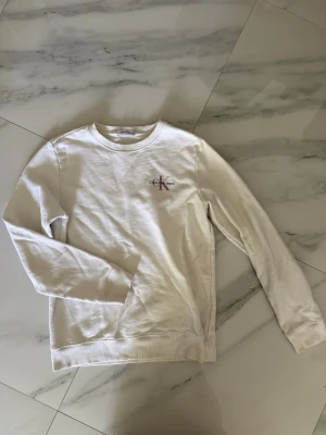 Vit sweatshirt från Calvin Klein - Säljer en stilren vit sweatshirt från Calvin Klein med diskret logga i rosa och svart på bröstet. Tröjan har rund halsringning, långa ärmar och ribbade muddar vid ärmslut och nederkant. Barnstorlek 170 men passar XS/S! 