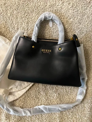 Svart handväska från Guess - Stilren svart handväska från Guess med guldfärgade detaljer och logga framtill. Väskan har både handtag och axelrem, samt flera fack inuti med dragkedja och ljusbeige foder med mönster. Perfekt för att bära dina viktigaste grejer med stil.