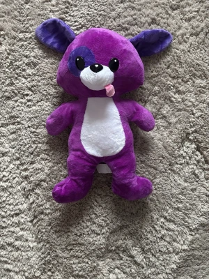 Lila gosedjur med stora öron - Mjukisdjur i lila plysch med stora öron och vitt parti på magen. Har ett lila fläck runt ena ögat, svart nos och en liten rosa tunga som sticker ut. Ögonen är svarta och blanka, och materialet är mjukt och gosigt. Perfekt som dekoration eller kramkompis.