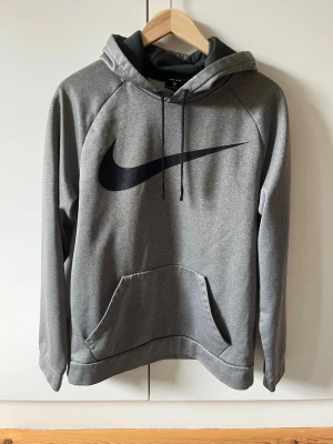 Grå hoodie Nike strl M - Snygg grå hoodie från Nike med stor svart logga på bröstet. Tröjan har huva med dragsko och en klassisk magficka. Material 100% polyester. Storlek M.