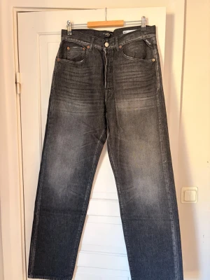 Svarta raka jeans med vintage-look - Säljer ett par svarta jeans med rak passform och  vintage-tvätt. Storlek: W32 | L34 . Jeansen har fem fickor, knappgylf och klassiska kontrastsömmar. Materialet är kraftig denim som ger en cool och avslappnad stil.