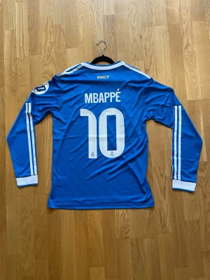 Mbappé - M - Säljer en blå långärmad Real Madrid matchtröja med Mbappé och nummer 10 på ryggen. Tröjan har vita detaljer, klubbmärke och Adidas-logga på bröstet samt Emirates Fly Better som sponsortryck. Tillverkad i ett lätt och ventilerande material, perfekt för fotbollsträning.
