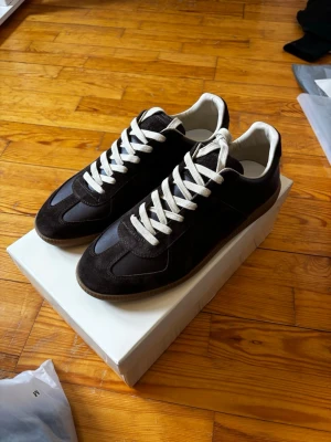 bruna MM sneakers med vita snören - Stilrena svarta sneakers med vita snören och beige sula. Skorna har en klassisk låg modell med ovandel i mocka och släta paneler i läder. Insidan är fodrad i vitt och designen är enkel men trendig, perfekt för en clean streetstyle-look.