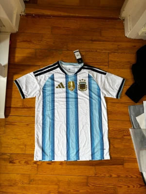 Argentina fotbollströja Adidas - Säljer en officiell Argentina fotbollströja från Adidas med klassiska blå och vita ränder. Tröjan har gulddetaljer, AFA-emblem och FIFA 2022-märke på bröstet. Tillverkad i polyester och perfekt för fotboll eller träning.