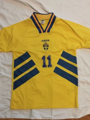 Svensk landslagströja Adidas nr 11 - Klassisk gul svensk landslagströja från Adidas med blå detaljer och nummer 11 på både fram- och baksida. Tröjan har BROLIN tryckt på ryggen, V-ringad krage med blå och gula ränder samt det svenska landslagsemblemet på bröstet. Perfekt för fotbollsfans!