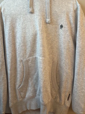 Grå hoodie från French Connection - Ljusgrå hoodie från French Connection med huva och dragsnören. Klassisk känguruficka framtill och liten svart logga på bröstet. Tillverkad i mjukt bomullsmaterial och har ribbade muddar vid ärmslut och nederkant. Perfekt för en chill och stilren look.