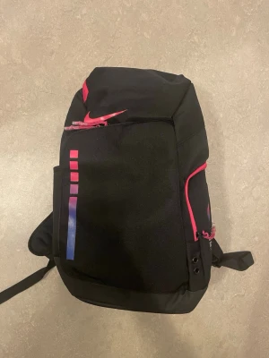 NIKE ELITE BACKPACK Svart ryggsäck från Nike med rosa detaljer - Snygg svart ryggsäck från Nike med rosa dragkedjor och logga. Framsidan har färgade rutor i rosa och blått. Axelremmarna är vadderade och har transparenta detaljer samt texten 'KAMPM'. Perfekt för skolan eller träningen.