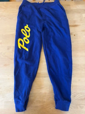 Blå ralph lauren mjukisbyxor - Snygga blå mjukisbyxor med stort gult Polo-tryck längs ena benet och gul bakficka. Byxorna har resår i midjan med dragsko och muddar vid bensluten. Perfekta för chill dagar eller när du vill ha en sportig look.