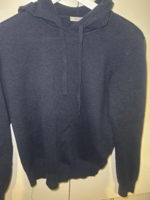 Mörkblå stickad hoodie med huva - Mysig mörkblå stickad hoodie med huva och dragsko. Tröjan har ribbade muddar vid ärmslut och nederkant. Perfekt för en avslappnad och stilren look.