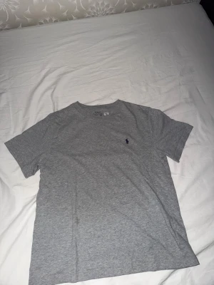 Grå t-shirt från Polo Ralph Lauren - Snygg grå t-shirt från Polo Ralph Lauren med klassisk rund hals och korta ärmar. Diskret logga broderad på bröstet i mörkblått. Tillverkad i mjuk bomull som känns skön mot huden. Perfekt basplagg till garderoben.