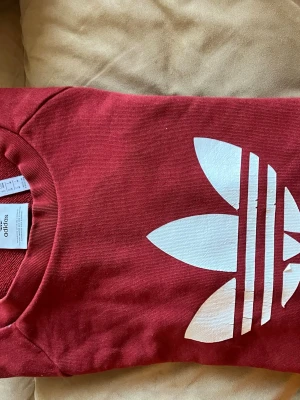 Röd sweatshirt från Adidas - Säljer en röd sweatshirt från Adidas med stort vitt Trefoil-tryck på bröstet. Klassisk rund halsringning och mjukt material som känns skönt mot huden. Perfekt för dig som gillar streetwear och sportig stil.