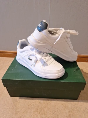 Vita Lacoste sneakers Game Advance - Fräscha vita junior sneakers från Lacoste, modell Game Advance. Skorna har klassisk låg siluett, perforerade detaljer på tån och sidan samt den ikoniska gröna krokodilloggan. Tillverkade i syntetmaterial med vita snören och diskret branding på hälen.