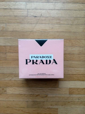 Oöppnad Prada Paradoxe Eau de Parfum 90ml - Prada Paradoxe Eau de Parfum i en elegant Ljusrosa kartong med svart detalj. Flaskan rymmer 90 ml och är en påfyllningsbar spray. Förpackningen har en modern och stilren design som känns lyxig och trendig. QR koden leder dig till pradabeauty's Instagram.