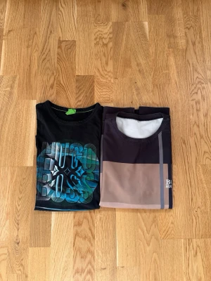 Två Hugo Boss t-shirts - Säljer två snygga t-shirts från Hugo Boss. Ena är svart med stort färgglatt tryck framtill, den andra har blockmönster i brunt, beige och svart med diskret logga på bröstet. Perfekta för en stilren och avslappnad look.