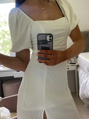 Vit långklänning med puffärm - Elegant vit långklänning med puffärmar och fyrkantig urringning. Klänningen har en slits framtill och är figurnära i modellen. Perfekt för sommarens alla tillfällen.