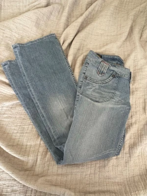 Ljusblå bootcut lågmidjade jeans med detaljer - Säljer ett par ljusblå lowwaist jeans med bootcut ben och coola sömdetaljer vid fickorna. Jeansen har klassisk femficksdesign och är gjorda i ett mjukt denimtyg. Från Lee i modellen LEOLA i storlek W29 L33
