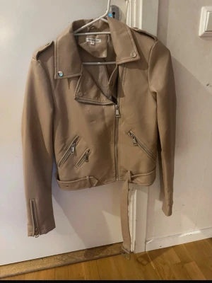 Beige skinnjacka med dragkedjor - Snygg beige skinnjacka med klassisk bikerstil, flera dragkedjeförsedda fickor och bälte i midjan. Jackan har axelklaffar, bred krage och dragkedjor vid ärmsluten för extra attityd. Perfekt för dig som vill ha en stilren och trendig look.