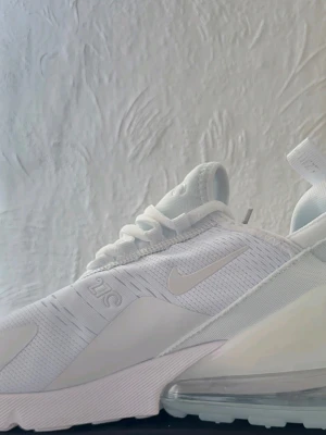 Nike Air Max 270 vita sneakers - Snygga vita Nike Air Max 270 sneakers med mesh- och syntetmaterial, tydlig Air Max-sula och diskret logga på sidan. Skorna har en sportig look med lågt skaft och stilrena detaljer. Perfekta för dig som gillar en clean och modern stil.