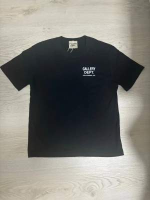 Svart Gallery Dept. t-shirt - Snygg svart t-shirt från Gallery Dept. med vit text på bröstet och stor logga på ryggen. Klassisk passform och rund hals. Perfekt för dig som gillar streetwear och enkel stil.