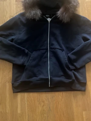 Svart hoodie med fuskpäls på huvan - Säljer en svart hoodie med dragkedja och stor huva kantad med fuskpäls. Hoodien har två stora fickor framtill och ribbade muddar vid ärmslut och nederkant. Perfekt för dig som vill ha en stilren men ändå lite extra hoodie.