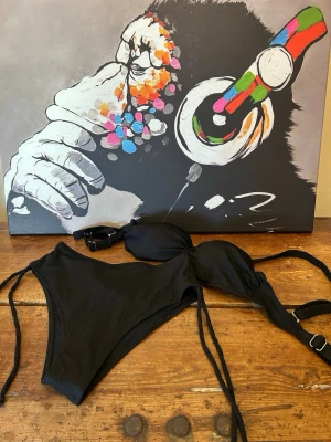 Svart bikini med snörning - Snygg svart bikini med knyt i sidorna och justerbara axelband. Bikinin har en diskret ribbad struktur och är tillverkad i mjuk polyesterblandning med elastan för skön passform. Perfekt för lata dagar på stranden eller poolen. Skyddsfilm finns kvar, helt ny. Aldrig använda 