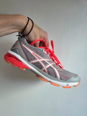 Asics GT 5000 - Snygga sneakers från Asics i grå mesh med vita och orange detaljer samt orange snören. Skorna har en sportig design med låg profil och en bekväm sula i vitt och orange. Perfekta för dig som gillar en färgstark och modern look.