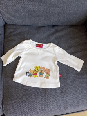 Vit långärmad Bamse t-shirt Lindex - Supersöt vit långärmad t-shirt från Lindex med Bamse-tryck framtill. Motivet visar Bamse och hans vänner i färgglada kläder. Tillverkad i mjuk bomull, perfekt för små barn. Rund halsringning och bekväm passform.