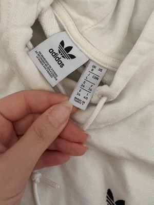 Vit hoodie från Adidas med svarta detaljer - Säljer en vit hoodie från Adidas med klassiska svarta ränder på axeln och broderad logga på bröstet. Hoodien har dragsko i huvan och är gjord i mjukt material, perfekt för en chill och sportig look.