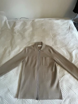 Beige overshirt från Samsøe Samsøe - Stilren beige overshirt från Samsøe Samsøe med dragkedja framtill, klassisk krage och en bröstficka. Tillverkad i ett mjukt, fleece-liknande material som ger en clean och modern look. Perfekt att slänga över en t-shirt för en chill vibe.