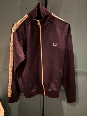 Mörk blå track jacket från Fred Perry - Säljer en snygg mörk blå track jacket från Fred Perry med beige detaljer och dragkedja framtill. Jackan har ribbade muddar, två fickor och beige band med Fred Perry-loggor längs ärmarna. Perfekt för en sportig och trendig look.
