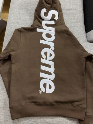 Brun Supreme hoodie - Säljer en brun hoodie från Supreme med stor vit logga längs ryggen. Hoodien har huva med dragsko och en klassisk känguruficka framtill. Tillverkad i mjukt bomullsmaterial och har ribbade muddar vid ärmslut och nederkant. Perfekt för dig som gillar streetwear.